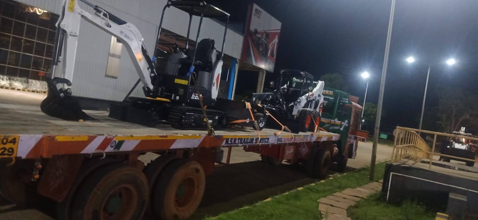 Compact excavator and skid-steer loader secured together on an MMM trailer under floodlights at night