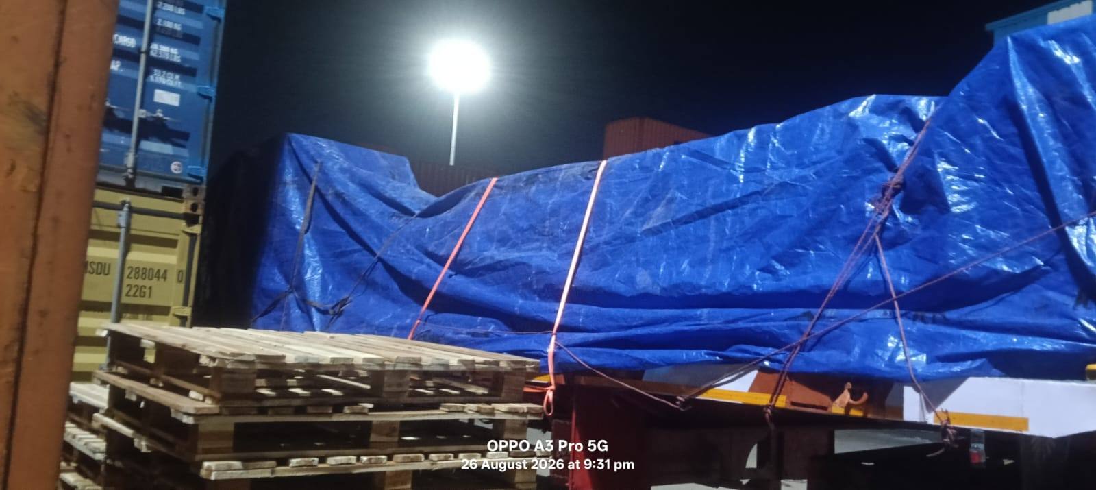 Blue tarpaulin covered load on a trailer under floodlights in a container yard